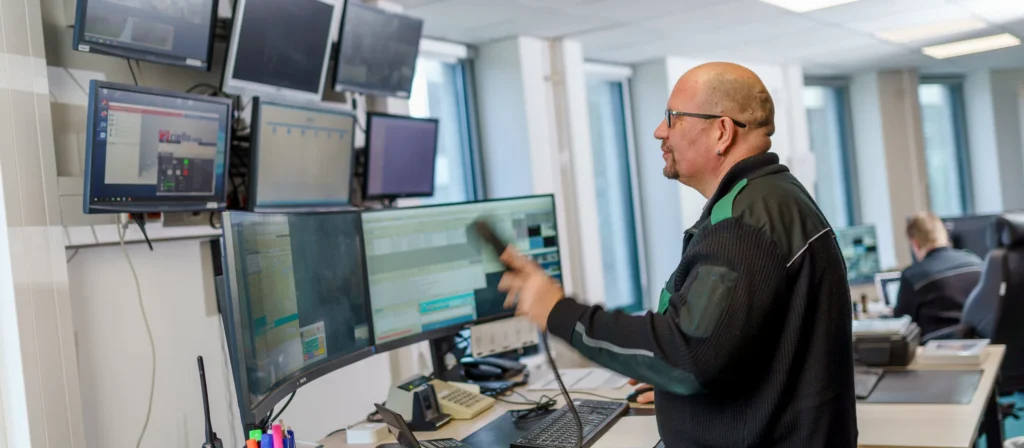 Ein Mann mit Brille steht in einer Leitstelle vor mehreren Monitoren und hält einen Telefonhörer; auf einem Bildschirm ist der Schriftzug Riello zu sehen.