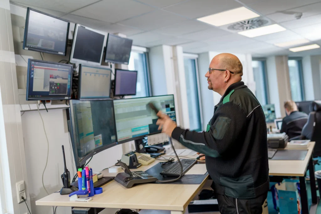 Ein Mann in dunkler Arbeitskleidung steht in einer Leitstelle vor mehreren Monitoren und hält einen Telefonhörer in der Hand.
