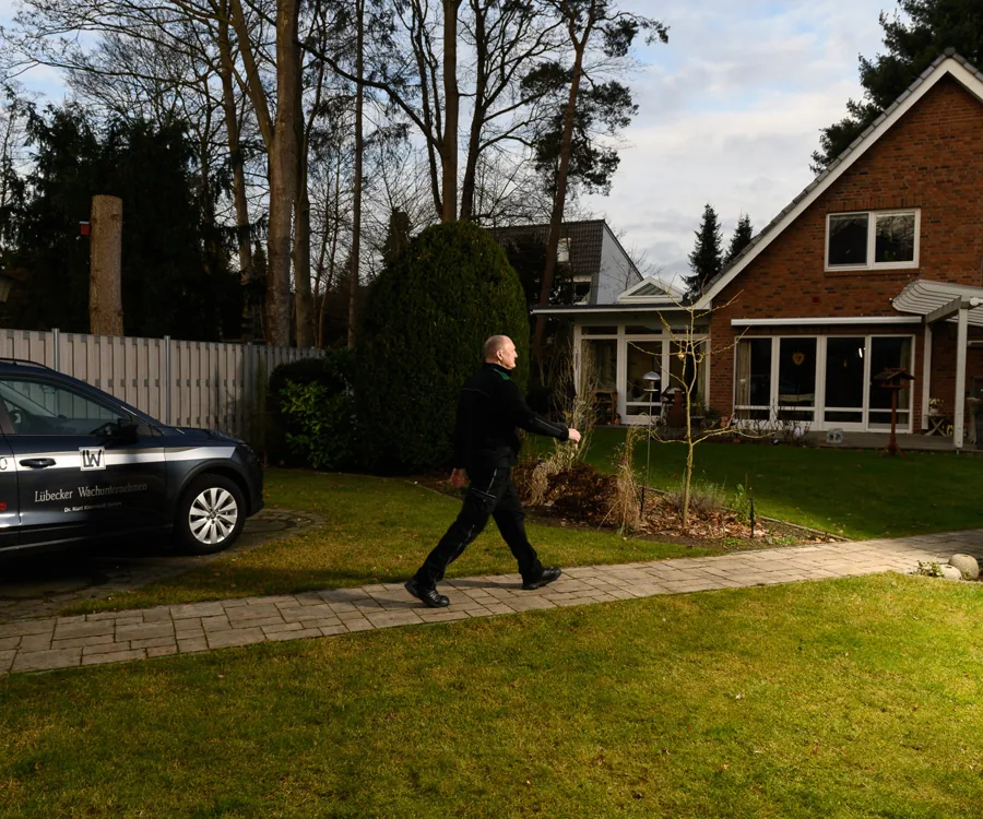 Ein Sicherheitsmitarbeiter eines Lübecker Wächterunternehmens geht auf einem Gartenweg, während sein Dienstfahrzeug daneben parkt.