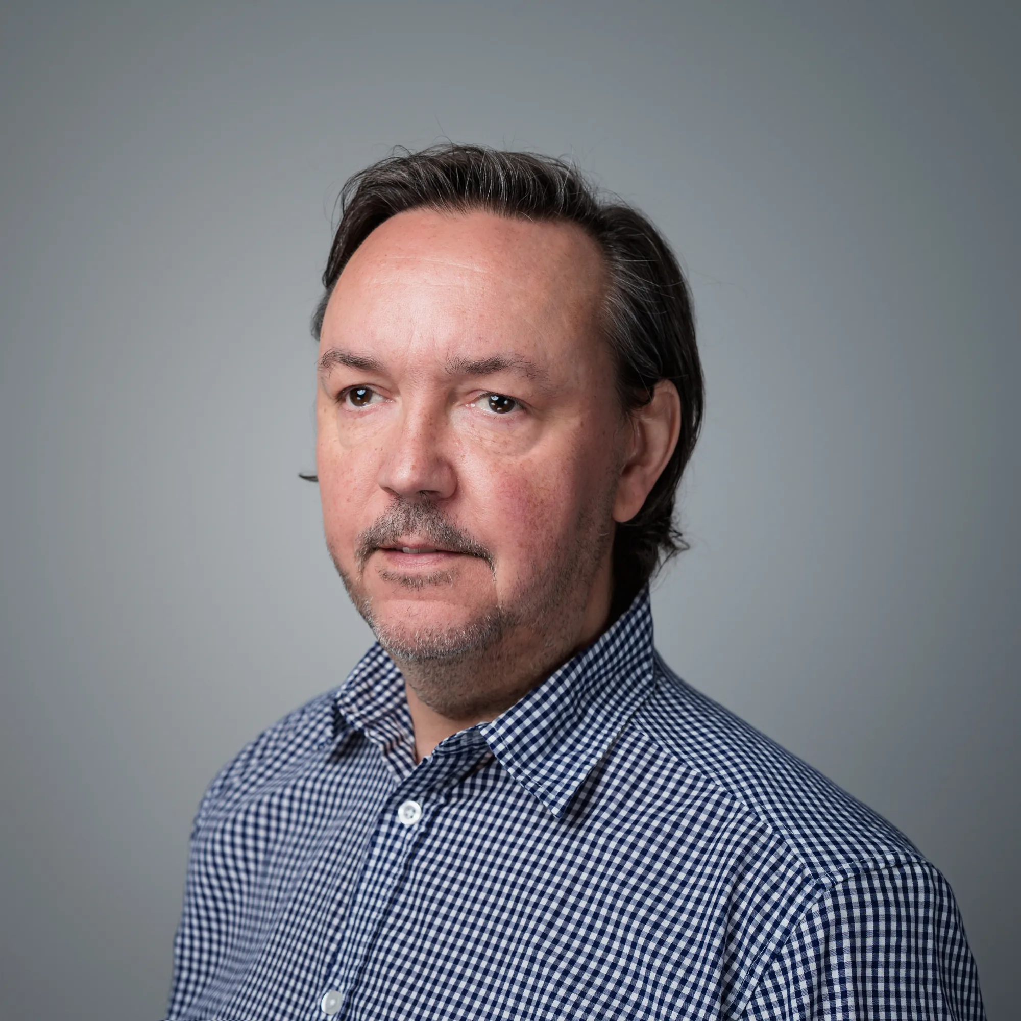 Porträt von Gerd Grzywatz, einem Mann mittleren Alters mit dunklem Haar und Bart, der ein blau-weiß kariertes Hemd trägt und zur Seite blickt.