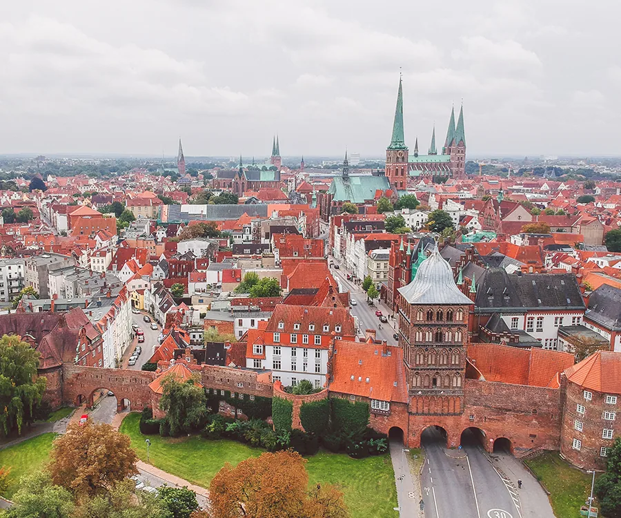 Luftaufnahme der historischen Altstadt von Lübeck mit dem Holstentor im Vordergrund und den markanten Kirchtürmen im Hintergrund unter bewölktem Himmel.