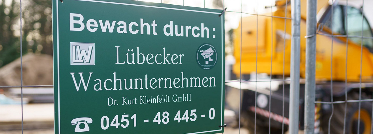 Grünes Schild „Bewacht durch Lübecker Wachunternehmen“ an einem Bauzaun.
