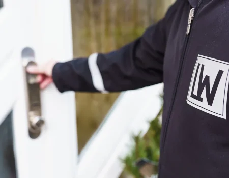 Eine Person in einer dunklen Jacke mit Firmenlogo öffnet eine weiße Tür, möglicherweise im Rahmen einer Alarmaufschaltung.