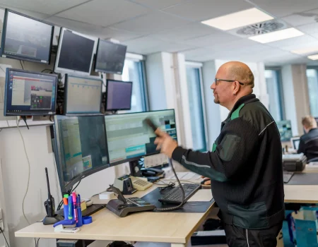 Ein Mann in Arbeitskleidung bedient mehrere Computermonitore und ein Kommunikationsgerät in einem Kontrollraum.