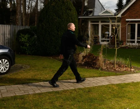 Ein Mann in dunkler Arbeitskleidung geht auf einem gepflasterten Weg durch einen Vorgarten, vorbei an einem Firmenwagen und einem Haus.
