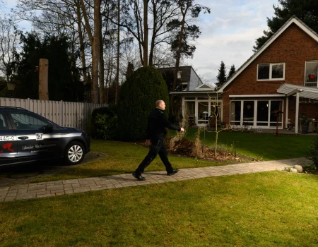 Ein Mann in dunkler Arbeitskleidung geht an einem geparkten Firmenwagen des Lübecker Wachunternehmens und einem Einfamilienhaus vorbei.