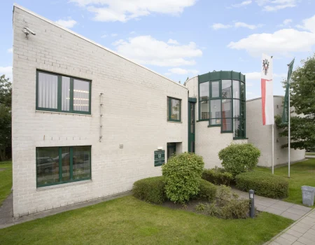 Ein modernes Gebäude mit hellen Ziegelwänden, grünen Fensterrahmen und einer runden Glasfassade, umgeben von Rasen und Büschen unter blauem Himmel.