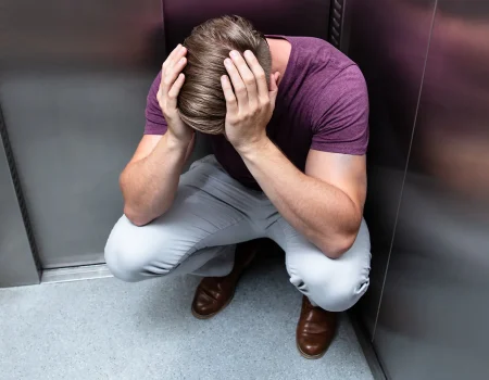 Ein Mann hockt verzweifelt in einem Aufzug, die Hände am Kopf, möglicherweise von Klaustrophobie oder einem Notfall betroffen.