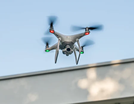 Eine weiße Kameradrohne fliegt vor einem hellen Himmel über einer Gebäudekante.